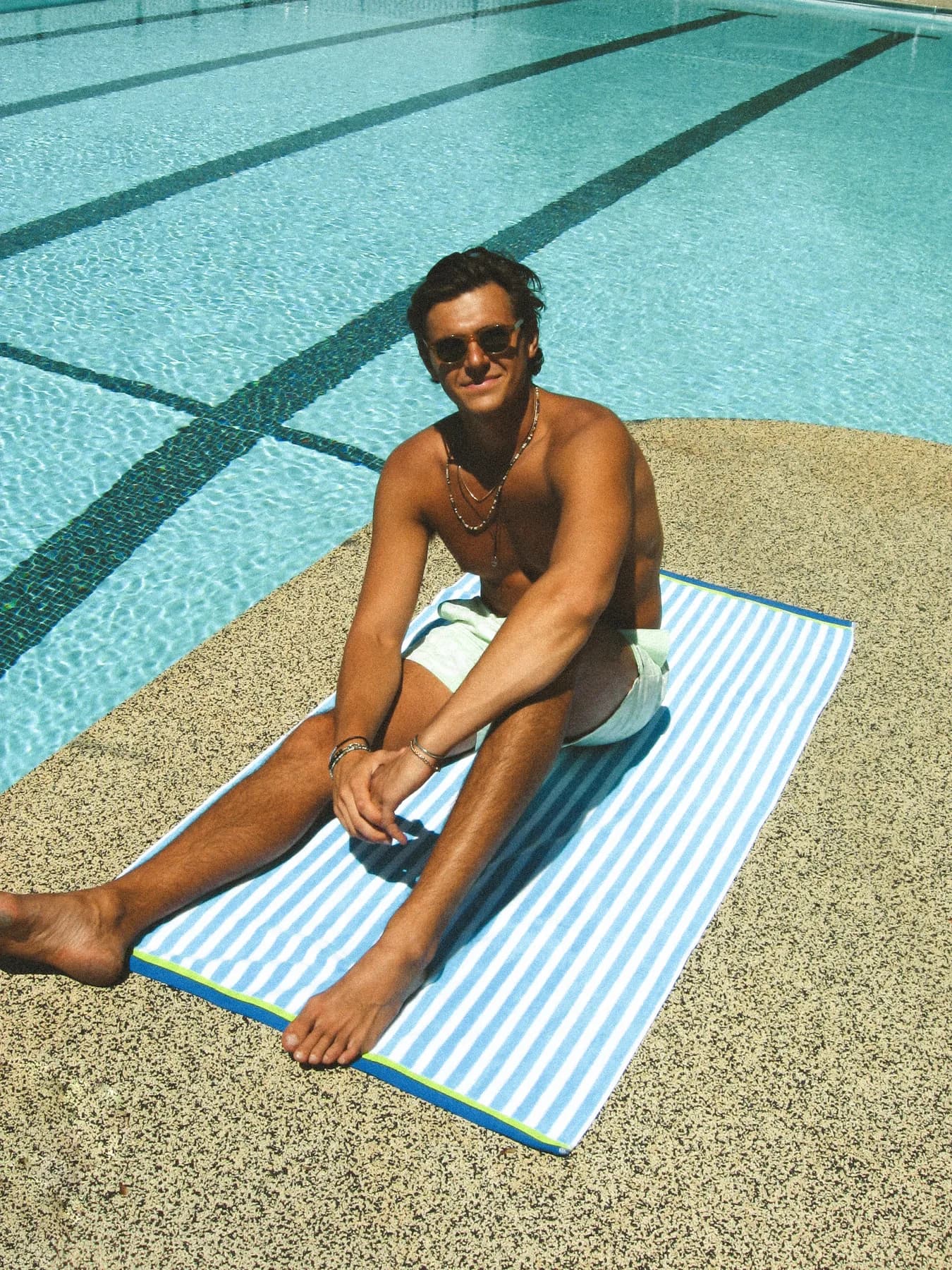 A person in sunglasses sitting on a striped towel beside a swimming pool